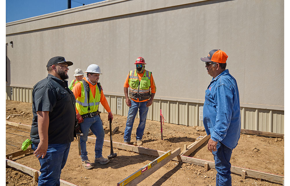 Five construction workers stand at a job site discussing plans near a foundation layout.
